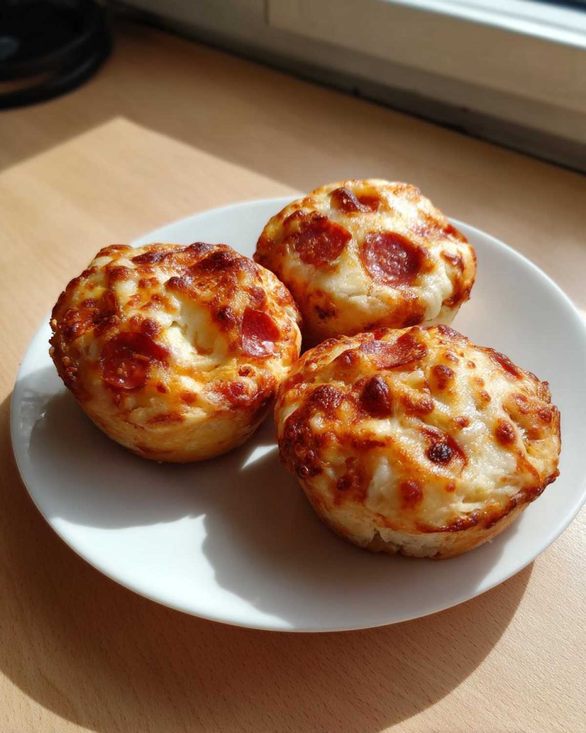 Three freshly baked pizza muffins topped with melted, browned cheese and pepperoni slices, served on a white plate.