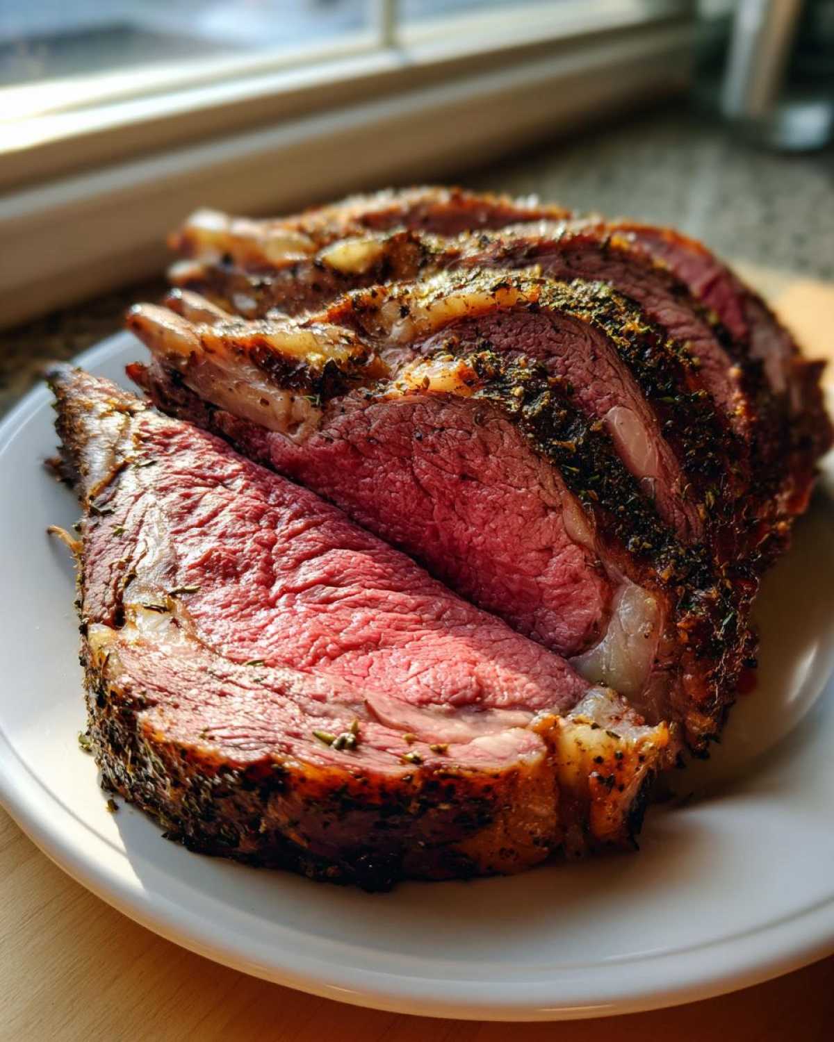 Several thick slices of medium-rare prime rib roast, crusted with herbs, resting on a white plate.
