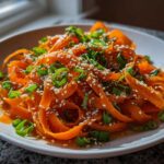 Close-up of a vibrant raw carrot salad made with shaved carrot ribbons, topped with sesame seeds and chopped green onions.