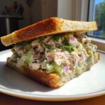 A thick, delicious salmon salad sandwich on toasted bread, filled with chunks of salmon, celery, and red onion, topped with fresh dill.