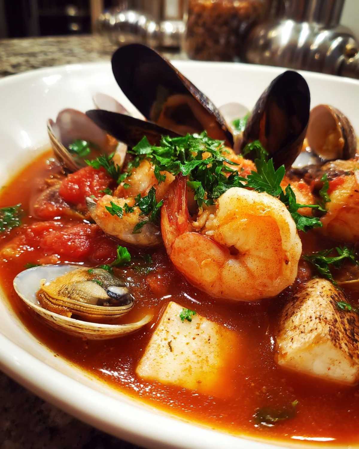Close-up of a rich, tomato-based seafood soup featuring shrimp, mussels, clams, fish chunks, and fresh parsley.