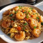 A close-up view of a white bowl filled with flavorful shrimp fried rice, topped with large shrimp, scrambled egg, and green onions.