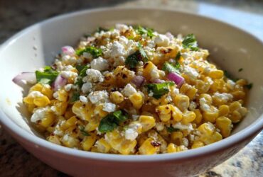 A close-up photo of a white bowl filled with vibrant yellow street corn salad topped with crumbled white cheese and fresh cilantro.