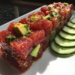 Close-up of glistening, marinated tuna tartare cubes topped with avocado and served alongside sliced cucumbers.