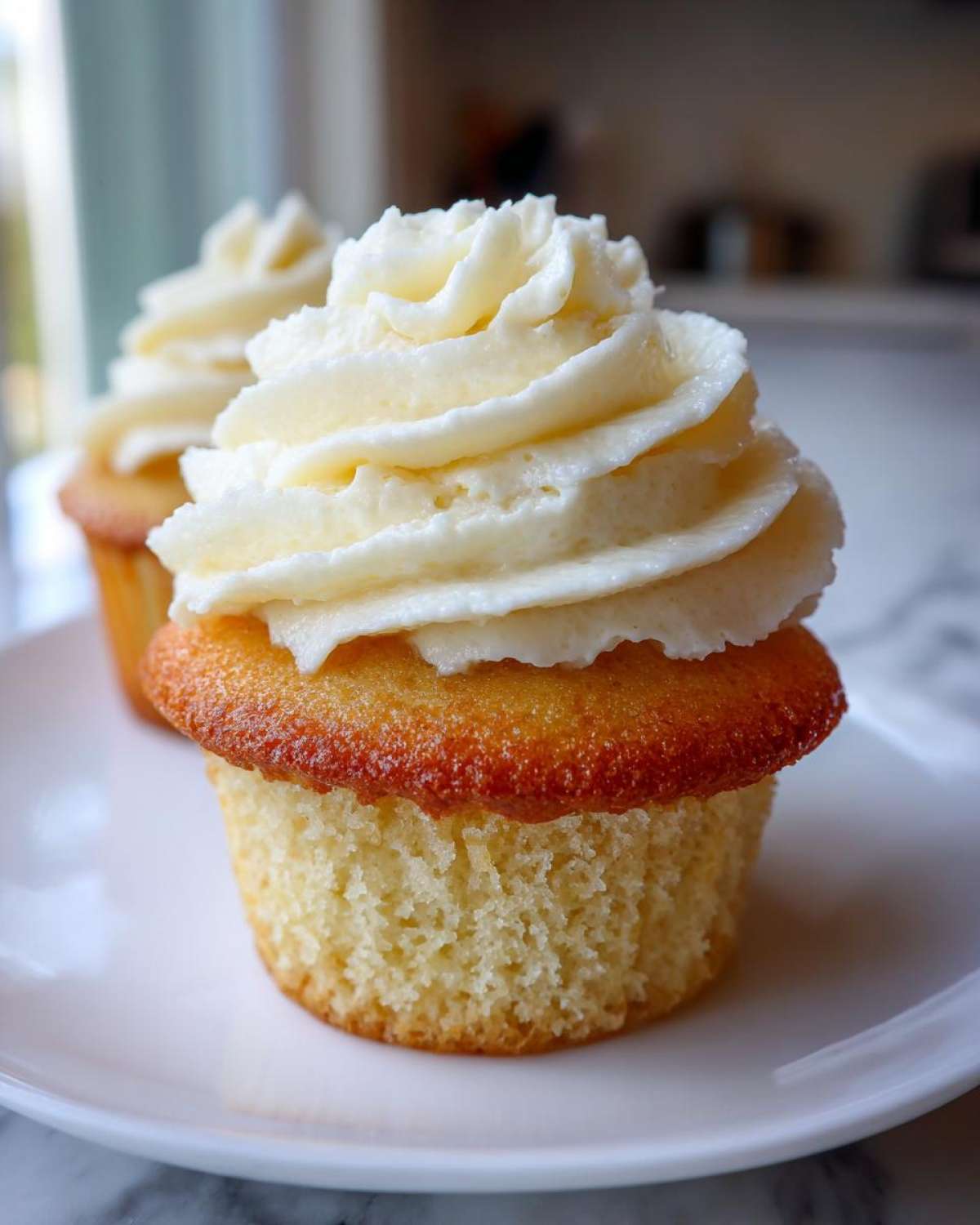 A close-up of an amazing vanilla cupcake topped with a swirl of white buttercream frosting, sitting on a white plate.
