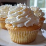Close-up of three amazing vanilla cupcakes topped with high swirls of creamy white frosting on a white plate.