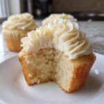Close-up of a vanilla cupcake with a bite taken out, revealing the moist crumb and creamy frosting of the amazing vanilla cupcakes.