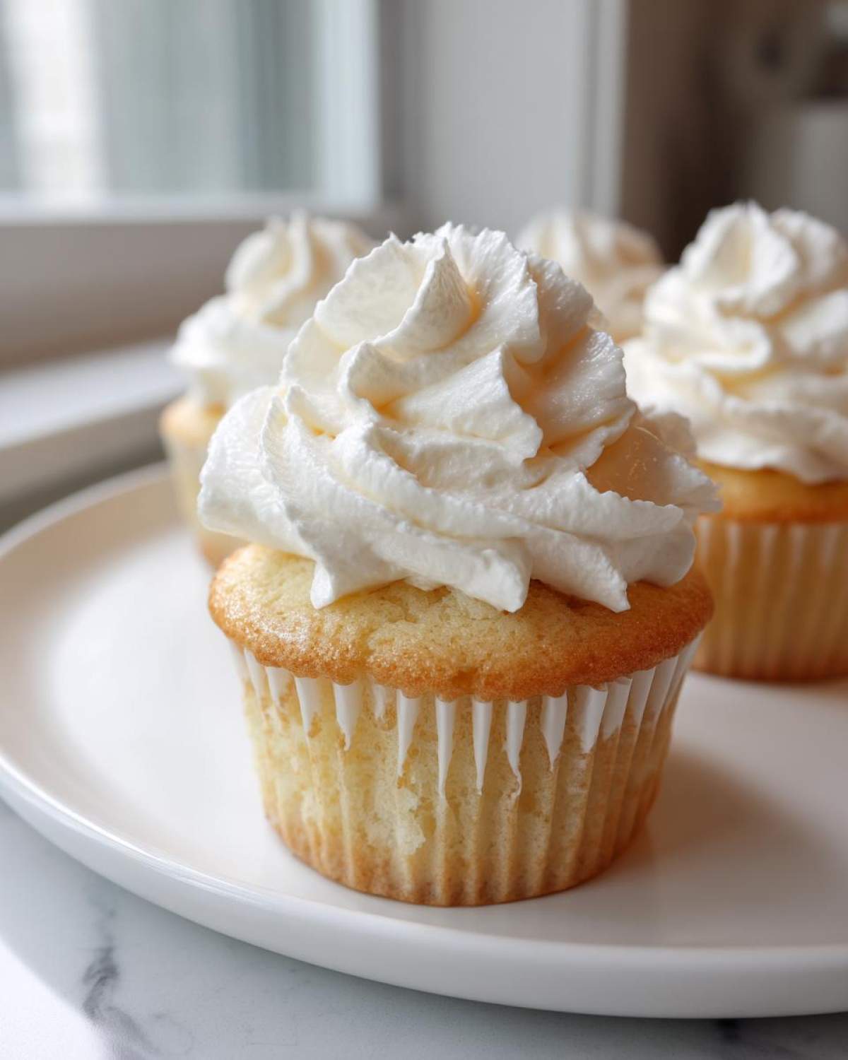 Close-up of a moist vanilla cupcake topped with a generous swirl of white frosting, sitting on a white plate.