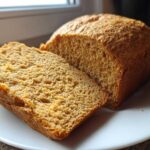Close-up of a golden brown whole wheat bread loaf with one thick slice cut and resting against it on a white plate.