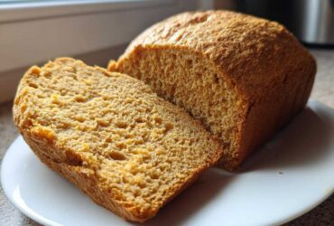 Close-up of a golden brown whole wheat bread loaf with one thick slice cut and resting against it on a white plate.