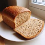 A freshly baked loaf of whole wheat bread, partially sliced, resting on a white plate near a window.