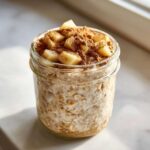 Close-up of apple cinnamon overnight oats in a glass jar, topped with diced apples and a generous dusting of cinnamon.