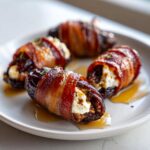 Four bacon wrapped stuffed dates filled with white cheese, drizzled with syrup on a white plate.