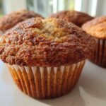 A close-up of a perfectly baked banana protein muffin with a golden-brown, textured top, sitting in a white liner.