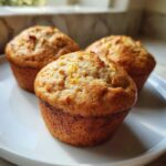 Three golden-brown banana protein muffins resting on a white plate, showcasing their textured tops.