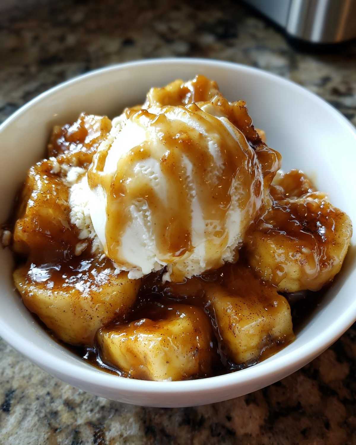 A white bowl filled with warm Bananas Foster, topped with a scoop of vanilla ice cream drizzled with caramel sauce.