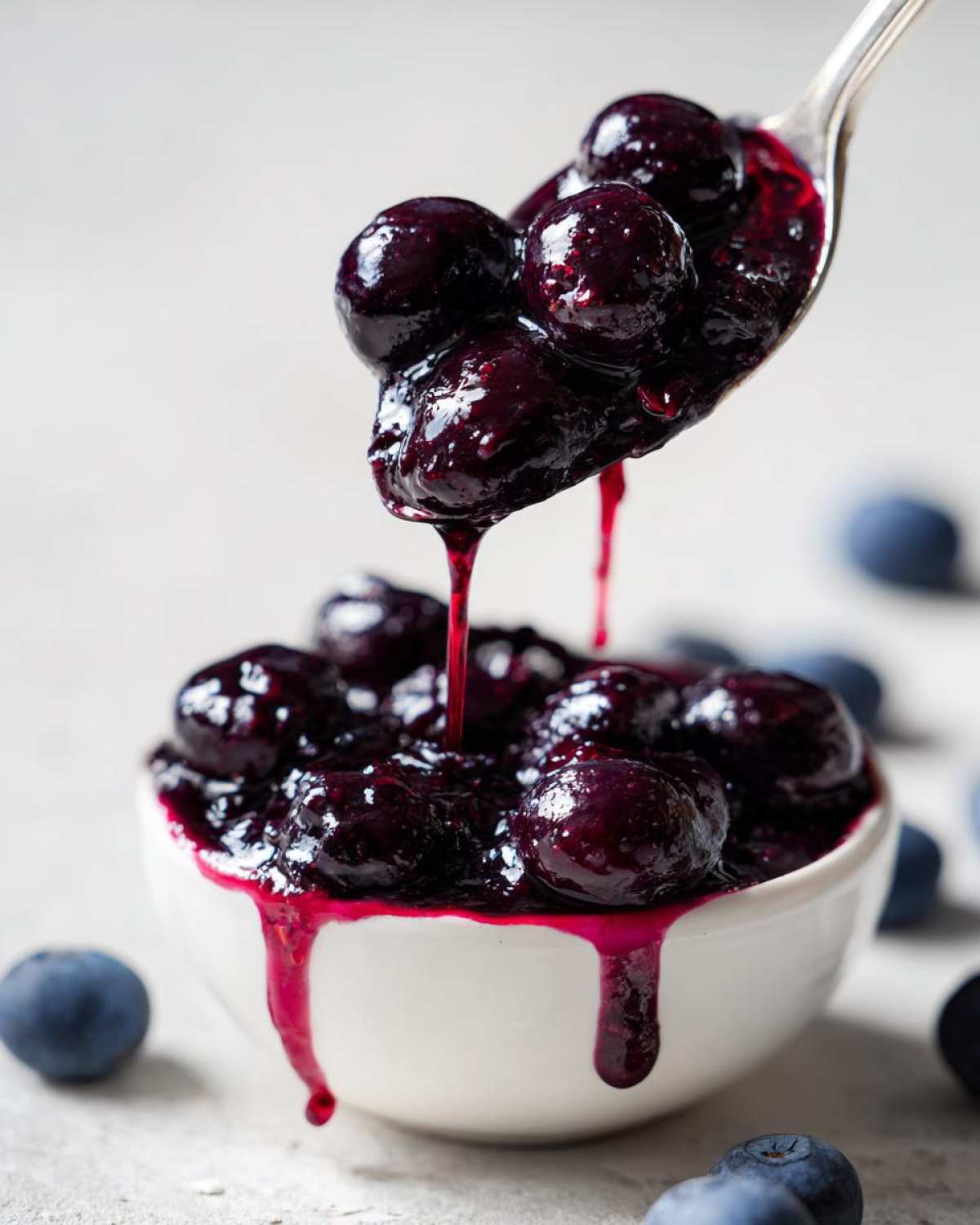 A spoonful of thick, glossy blueberry jam is lifted from a white bowl, dripping down the sides.