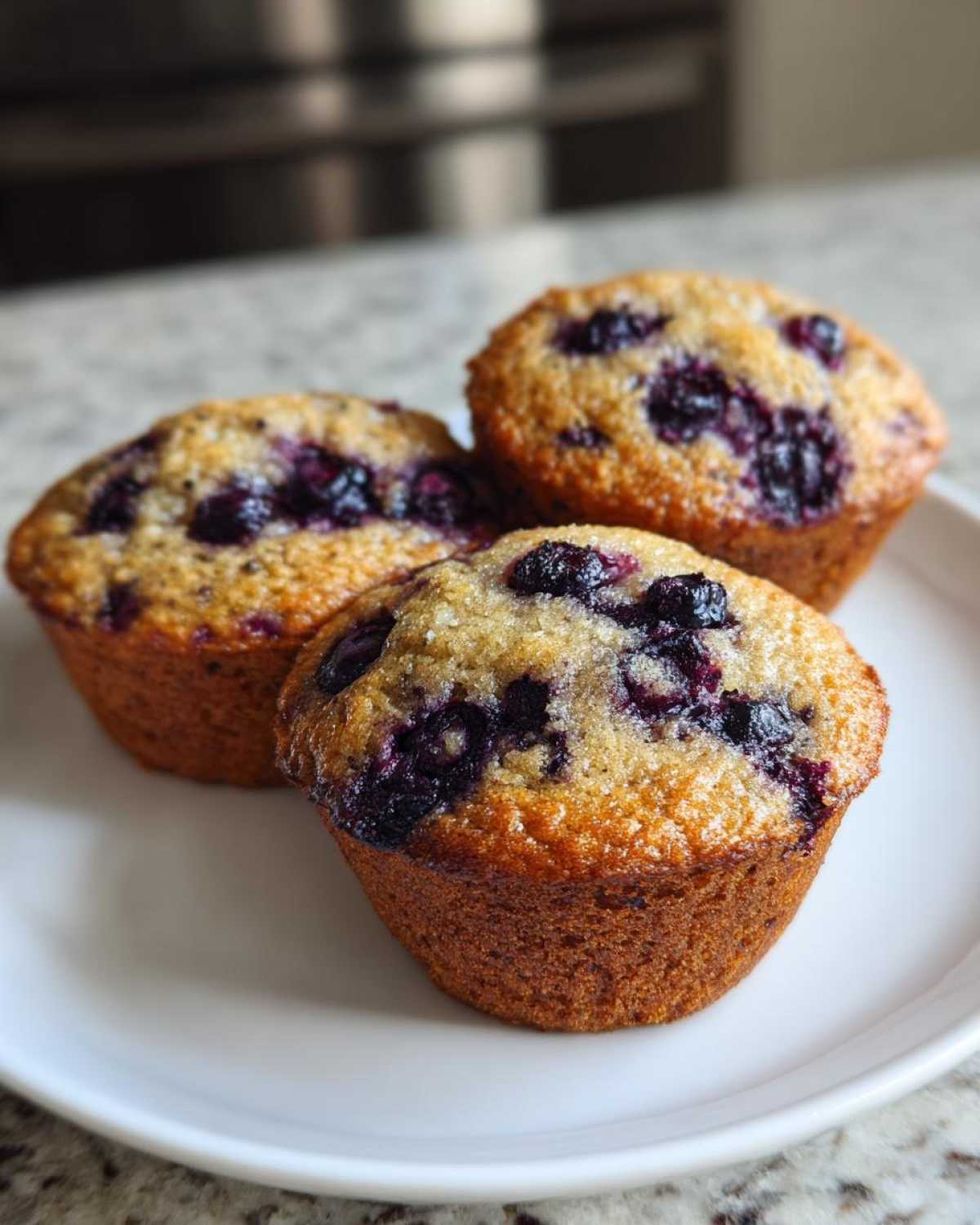 Three freshly baked blueberry protein muffins with a sugary top resting on a white plate.