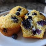 A plate featuring several moist blueberry protein muffins, one cut in half to show the crumb and fruit.