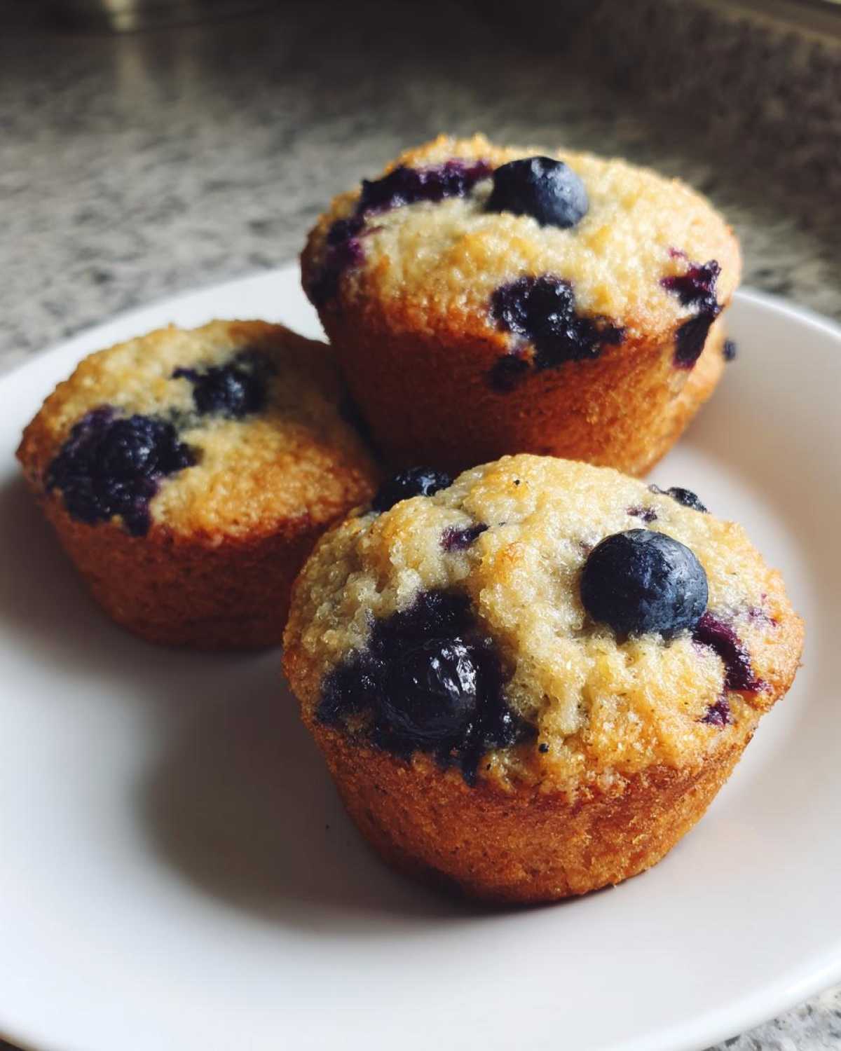 Three golden-brown blueberry protein muffins stacked closely on a white plate.