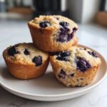 Three freshly baked blueberry protein muffins stacked slightly on a light-colored plate.