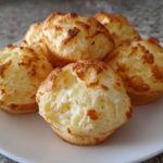 A close-up of several golden-brown Brazilian cheese bread puffs piled on a white plate.