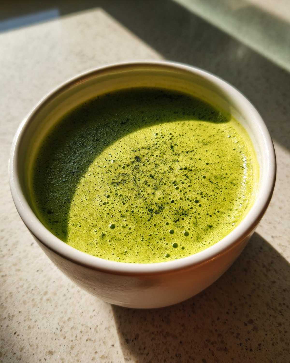 Close-up of a bright green, foamy drink, possibly matcha, in a white mug, contrasting with sunlight.