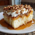 A decadent slice of butter pecan praline poke cake soaked in caramel sauce, topped with whipped cream and chopped pecans.
