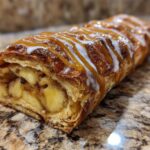 A close-up of a flaky pastry filled with cinnamon apples, drizzled with caramel and white icing, representing an apple strudel.