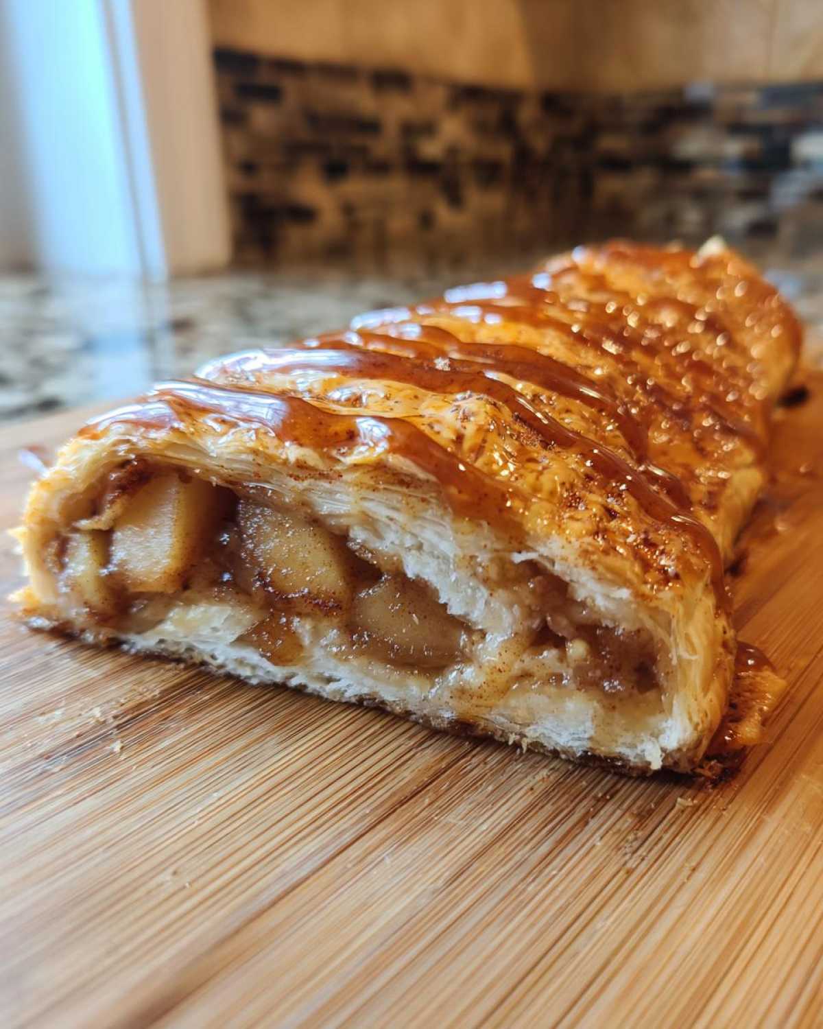 A close-up view of a freshly baked apple strudel slice, showing flaky pastry and caramelized apple filling, drizzled with caramel sauce.