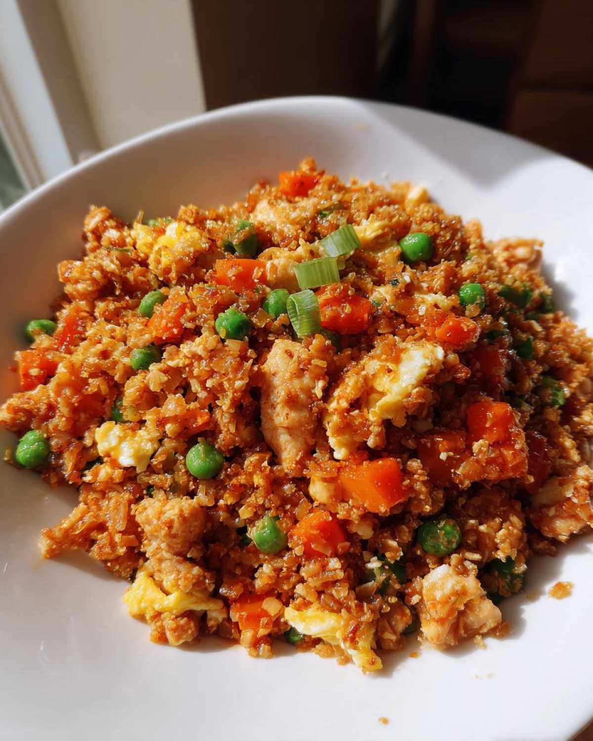 A close-up of a white bowl filled with savory cauliflower fried rice, mixed with scrambled egg, peas, and carrots.