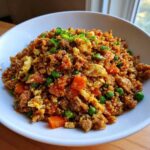 A close-up of a white bowl filled with savory cauliflower fried rice, mixed with scrambled egg, carrots, and green peas.