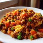 A close-up serving of vibrant cauliflower fried rice mixed with scrambled egg, bright green peas, and diced carrots.