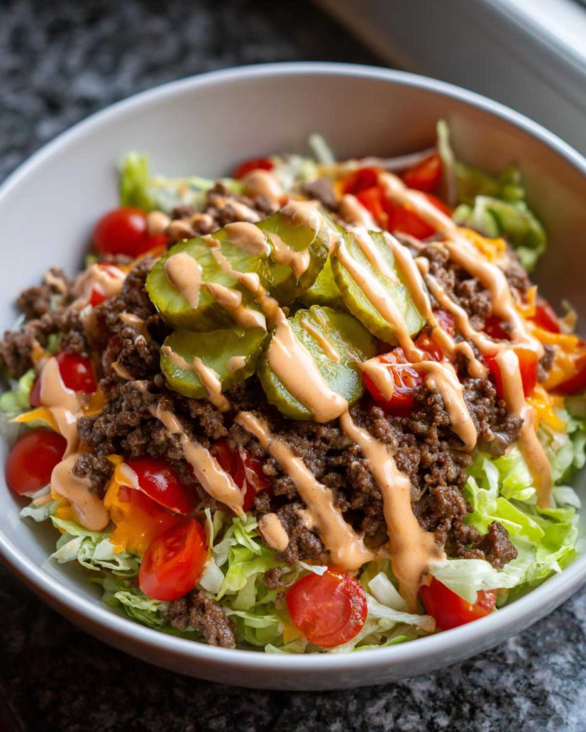 A close-up of a fresh cheeseburger bowl featuring seasoned ground beef, shredded lettuce, cherry tomatoes, pickles, and a drizzle of sauce.