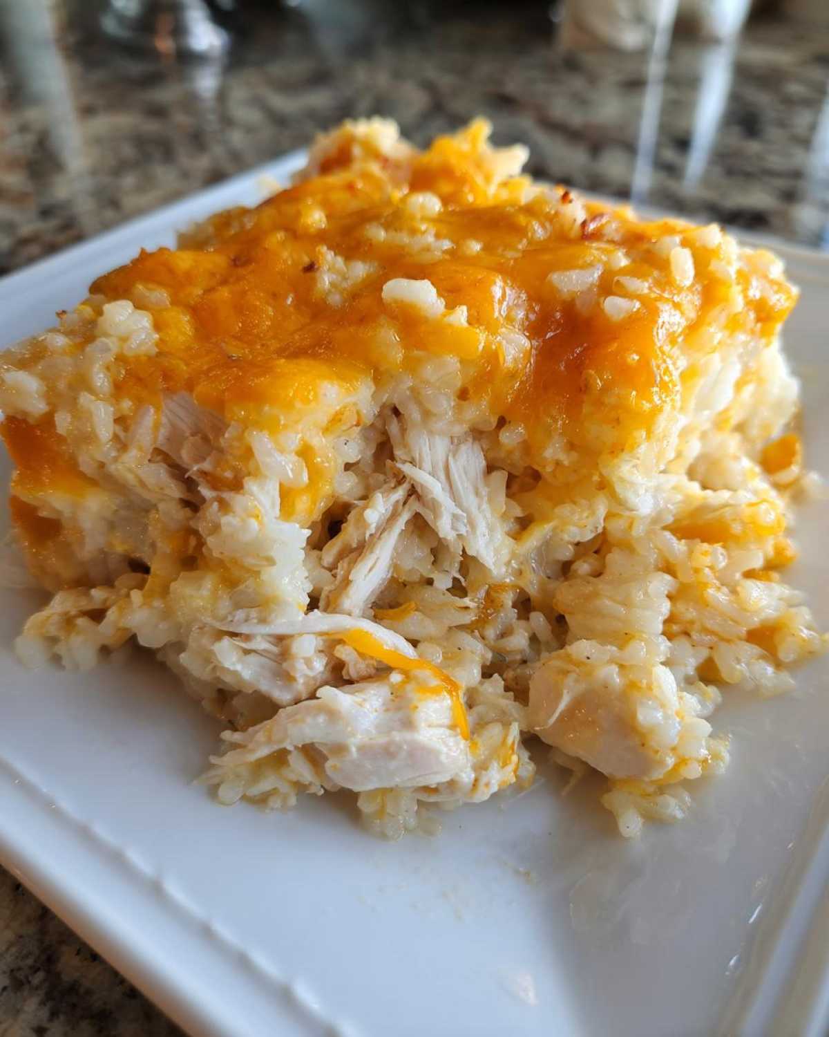 Close-up of a square serving of cheesy chicken and rice casserole topped with melted cheddar cheese.