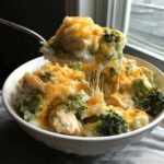 A spoonful of creamy chicken broccoli rice casserole showing a dramatic cheese pull over a white bowl.
