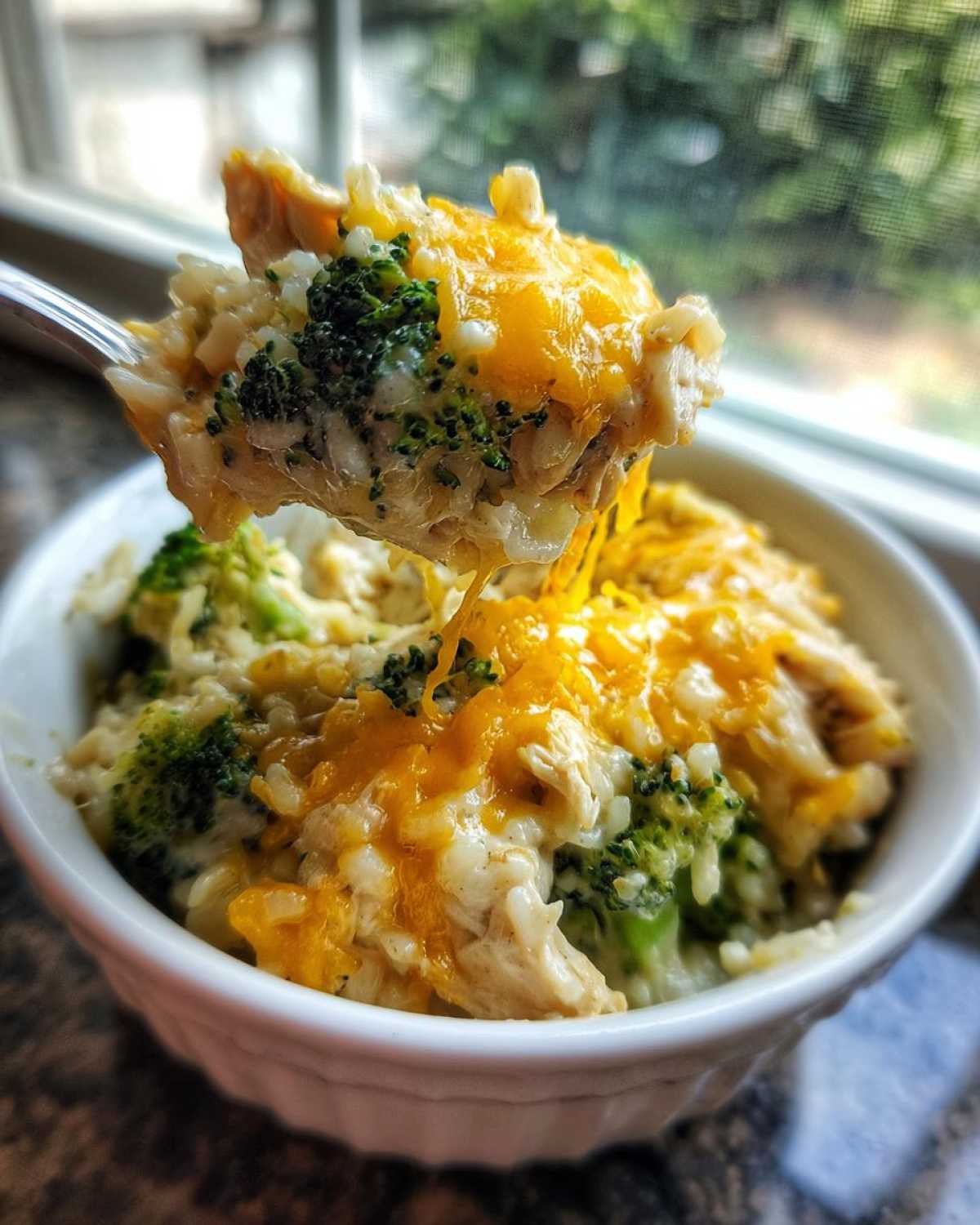 A spoonful of creamy chicken broccoli rice casserole topped with melted cheddar cheese being lifted from a white bowl.