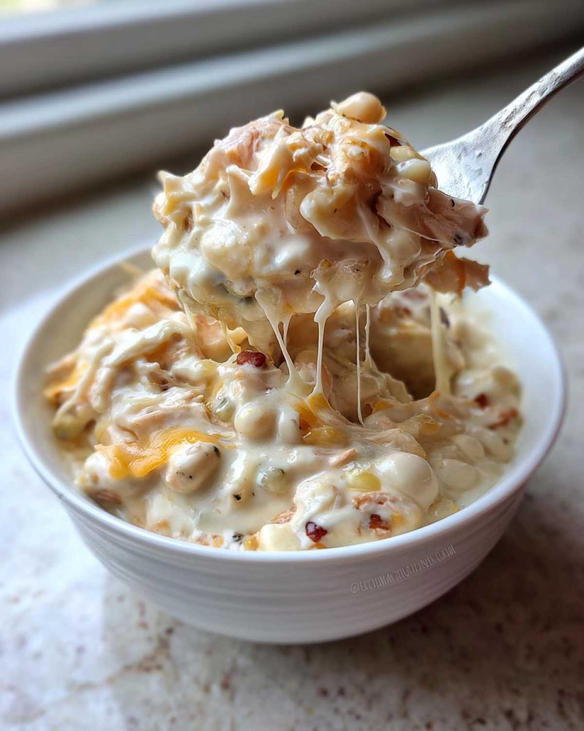A spoonful of creamy crockpot white chicken chili being lifted from a white bowl, showing melted cheese strings.