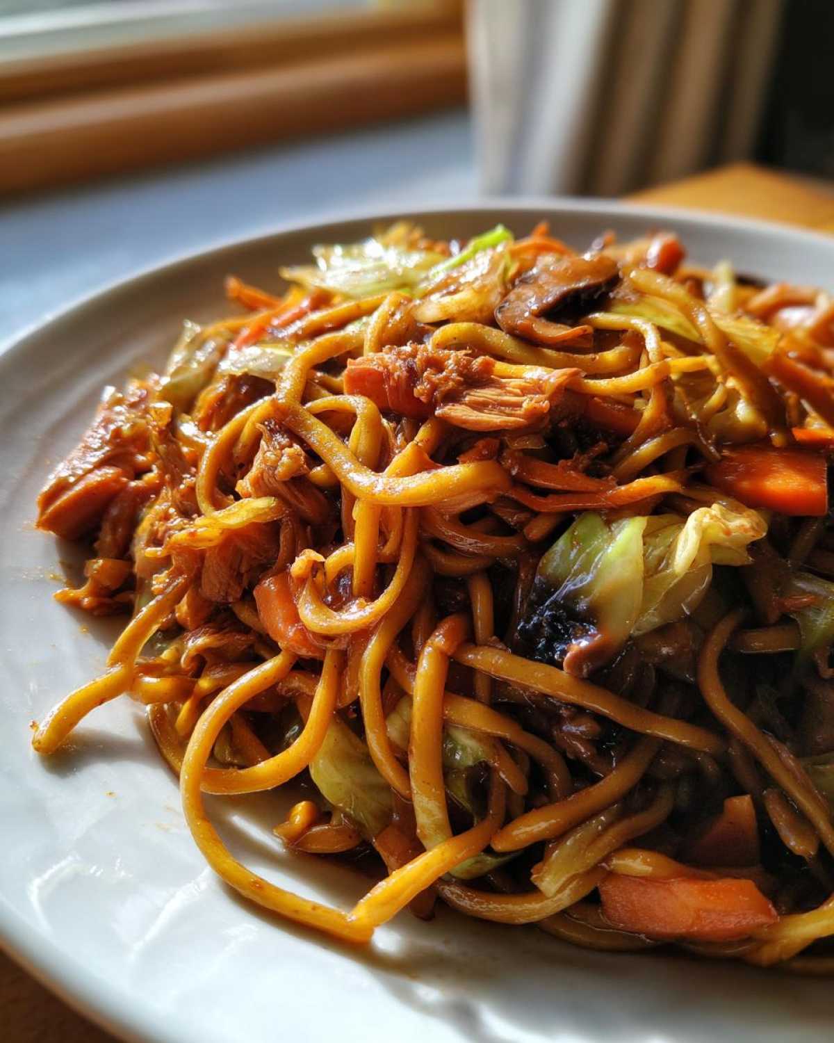 Close-up of a plate piled high with saucy chicken chow mein recipe featuring noodles, shredded chicken, cabbage, and carrots.
