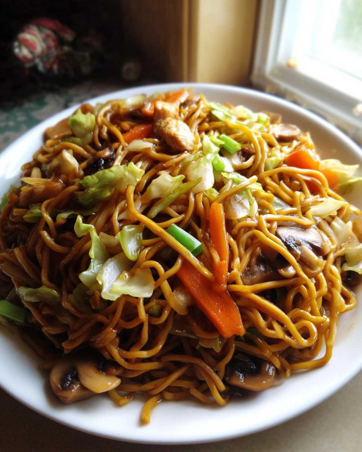 A close-up of a serving of homemade chicken chow mein recipe, featuring noodles, chicken pieces, carrots, cabbage, and mushrooms.