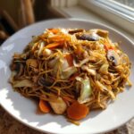 A close-up of a serving of chicken chow mein recipe, featuring noodles, chicken pieces, carrots, cabbage, and mushrooms.