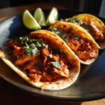 Three delicious chicken tinga tacos served on a dark plate with fresh cilantro and lime wedges.