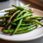 A close-up of bright green, blistered chinese green beans with garlic sauce piled on a white plate.