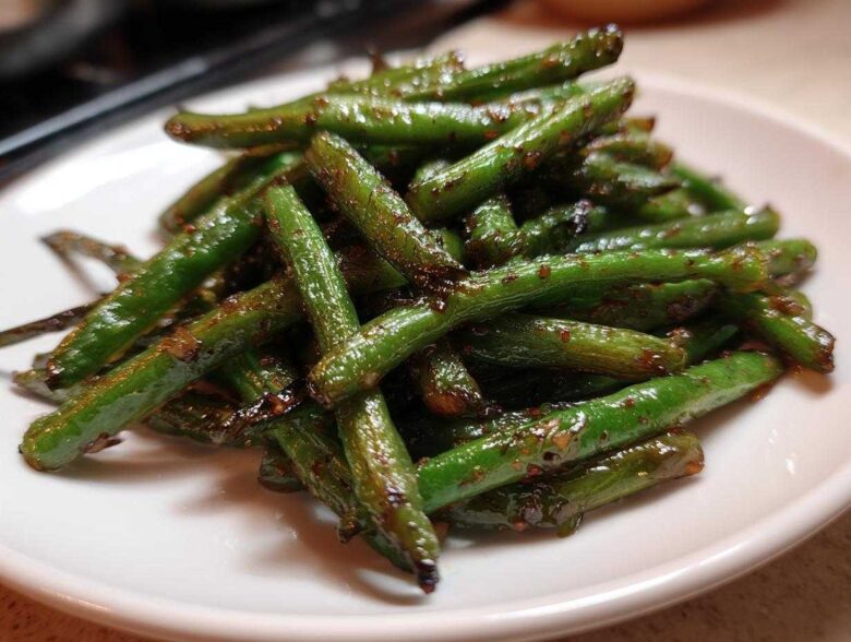 A pile of glossy, stir-fried chinese green beans with garlic sauce served on a white plate.