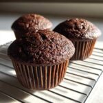 Three dark, rich chocolate bran muffins cooling on a wire rack in bright sunlight.