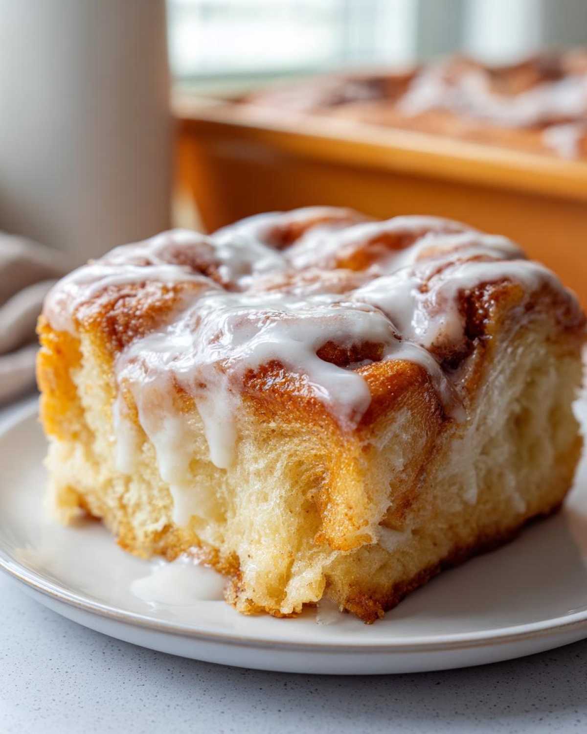 A close-up shot of a single, fluffy square serving of cinnamon roll casserole topped with thick white icing.