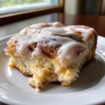 A close-up shot of a fluffy slice of cinnamon roll casserole topped with thick white icing.