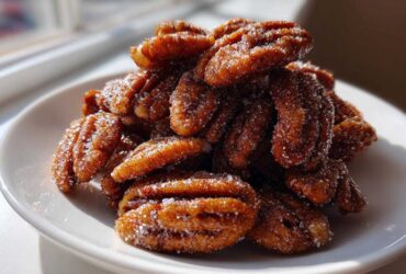 A pile of glistening cinnamon sugar pecans, coated in visible sugar crystals, served on a small white plate.