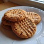 Close-up of four golden brown peanut butter cookies stacked on a white plate, showing classic crosshatch fork marks.