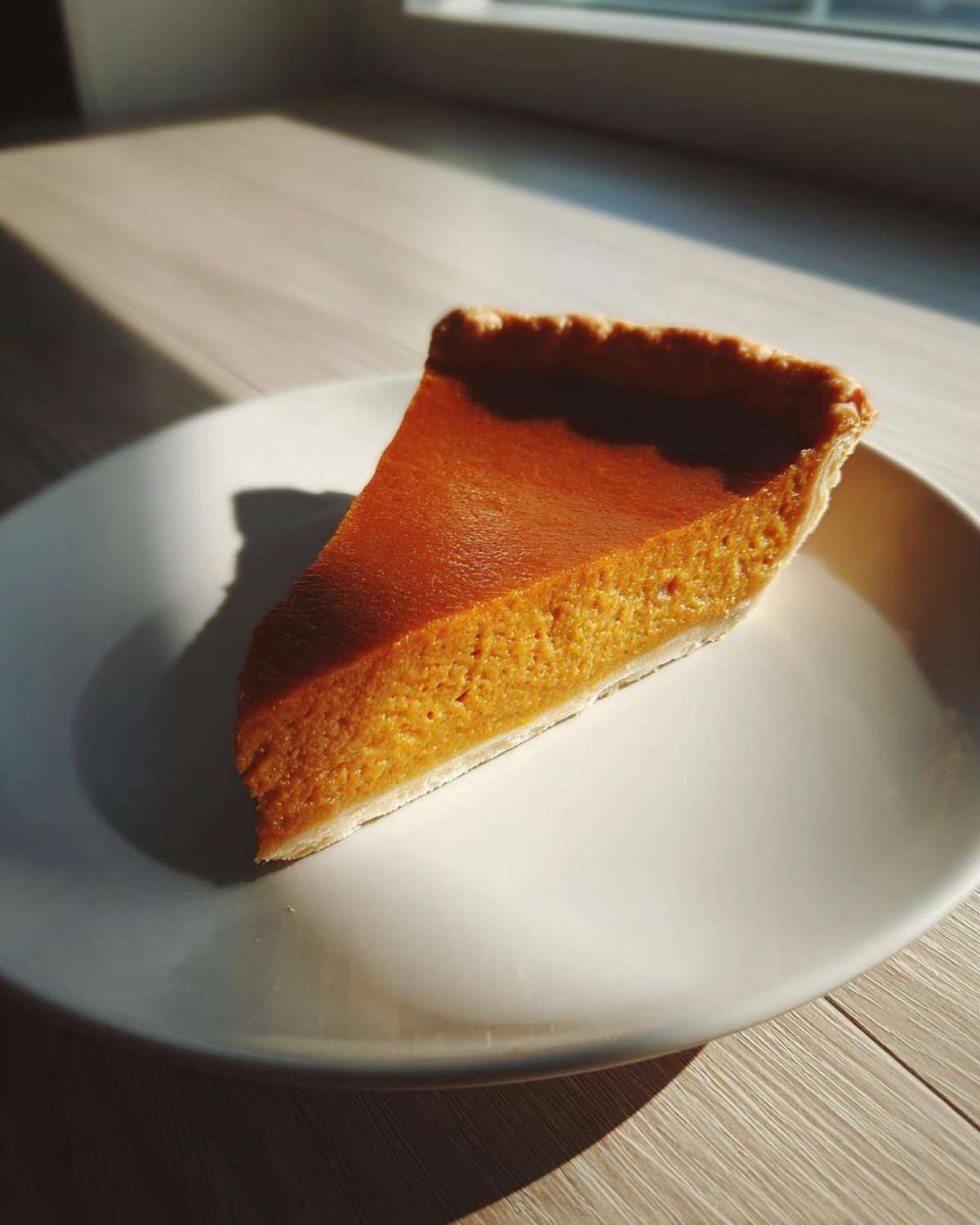 A perfect slice of classic pumpkin pie recipe sitting on a white plate, highlighted by dramatic sunlight.